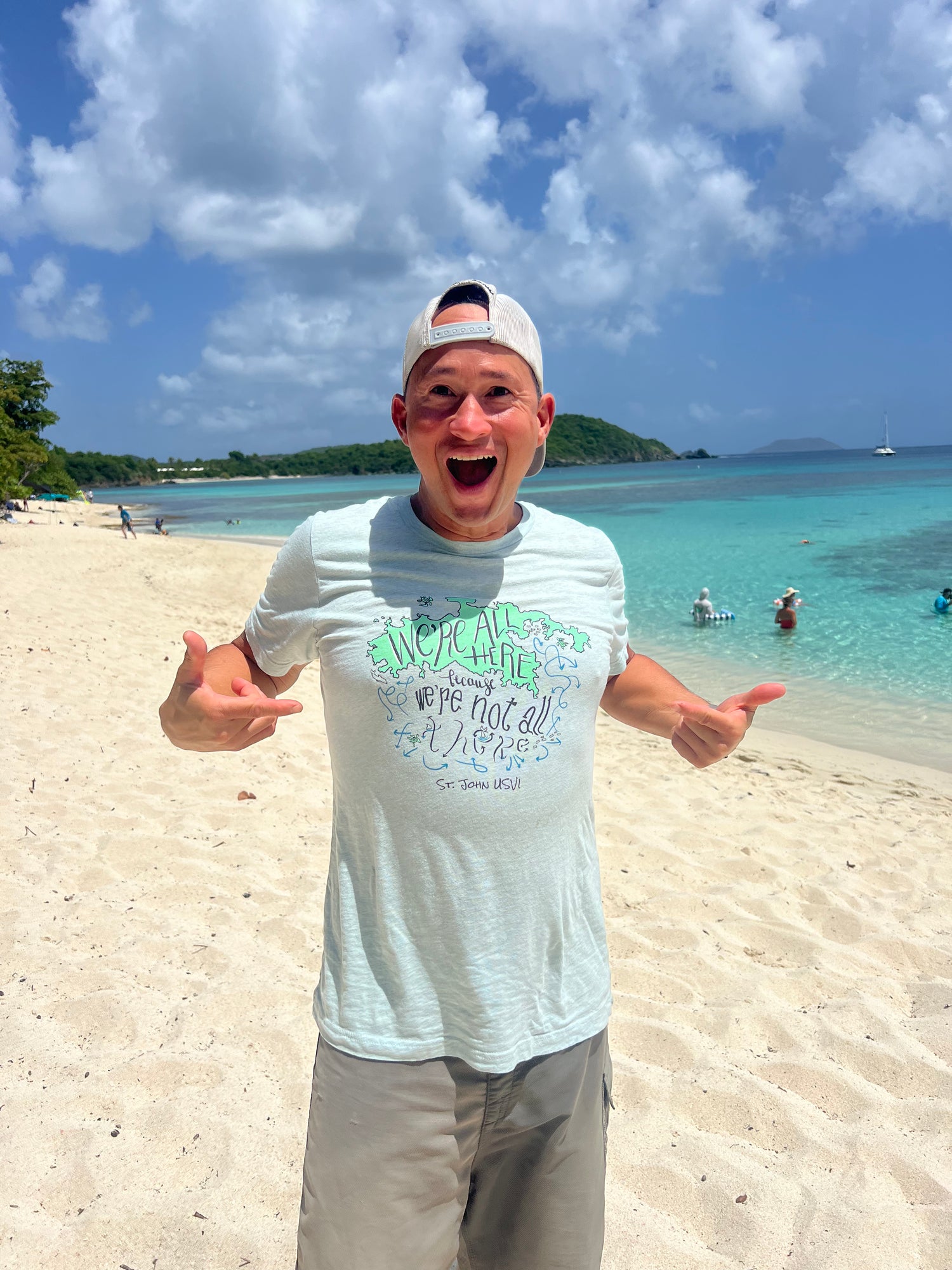 St. John USVI resident Mat excitedly wearing an original art souvenir t-shirt while standing on Hawk's Nest Beach with beautiful Caribbean ocean blue water in background